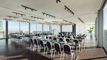 Uma sala de conferências ampla, com várias mesas e cadeiras, grandes janelas com vista para a cidade e um design de teto moderno com iluminação em trilhos.