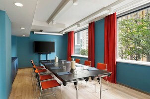 Una sala conferenze con un lungo tavolo, sedie, una TV a schermo piatto e una finestra con tende rosse.
