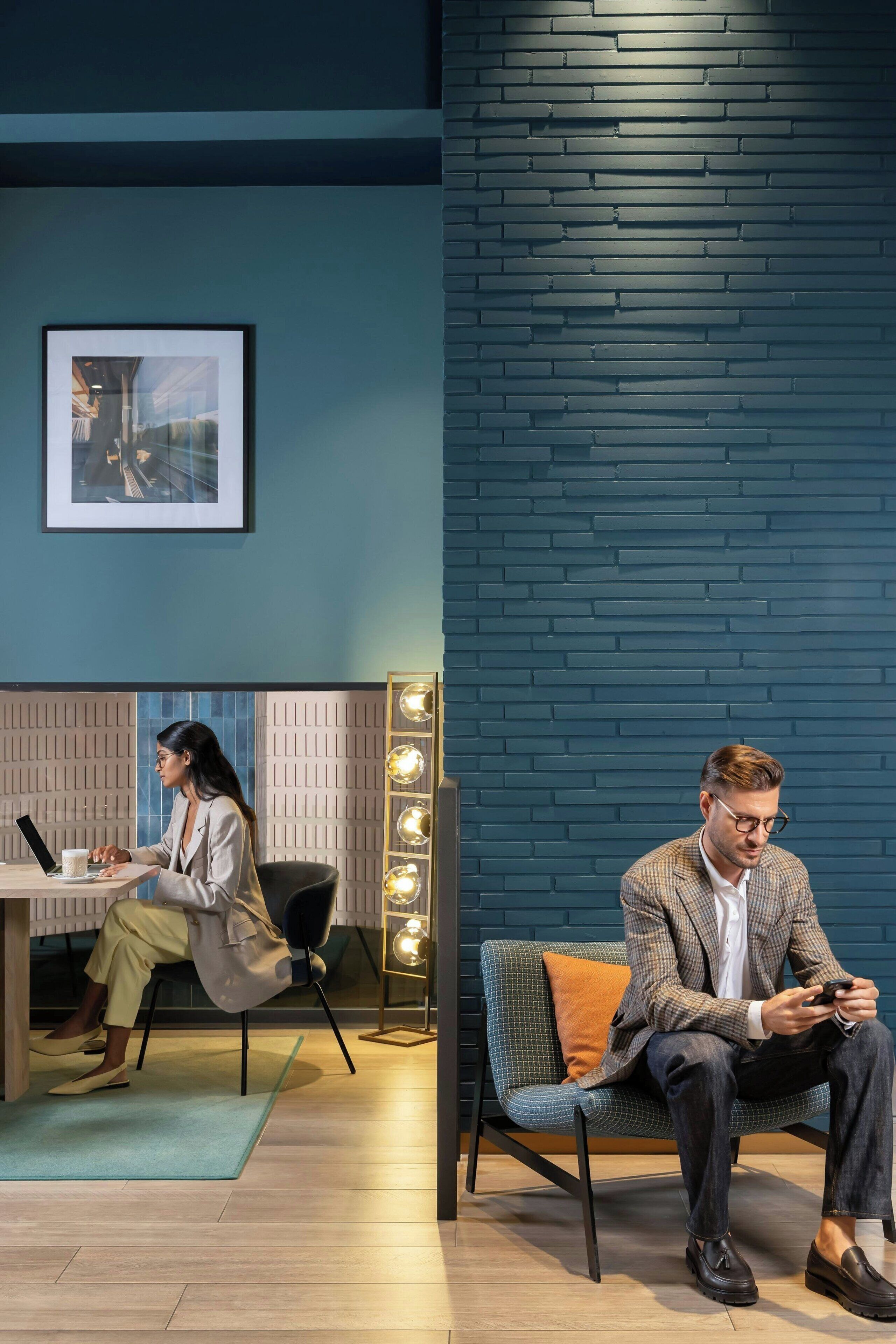 A modern room with a blue brick wall, a framed picture, a desk with a laptop, and a seating area with a blue chair and a lamp.