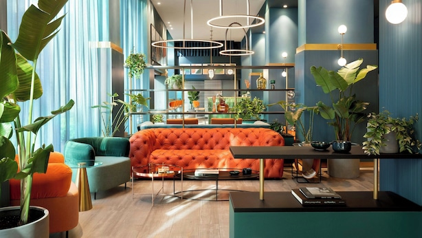 A modern living room with a tufted orange sofa, teal armchairs, and a coffee table. There are multiple plants, a bookshelf, and a dining area visible. - Duo Milan Porta Nuova, A Tribute Portfolio Hotel (Milan)