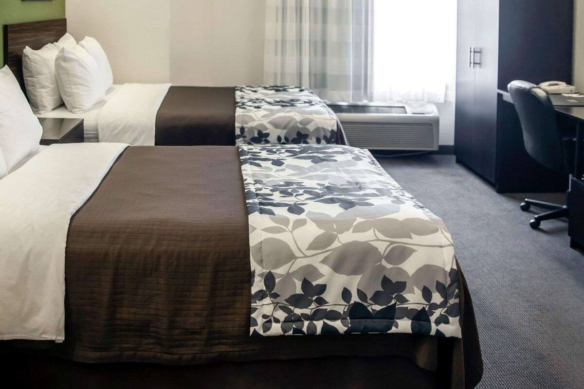 A hotel room with two beds, a desk, and an air conditioning unit.
