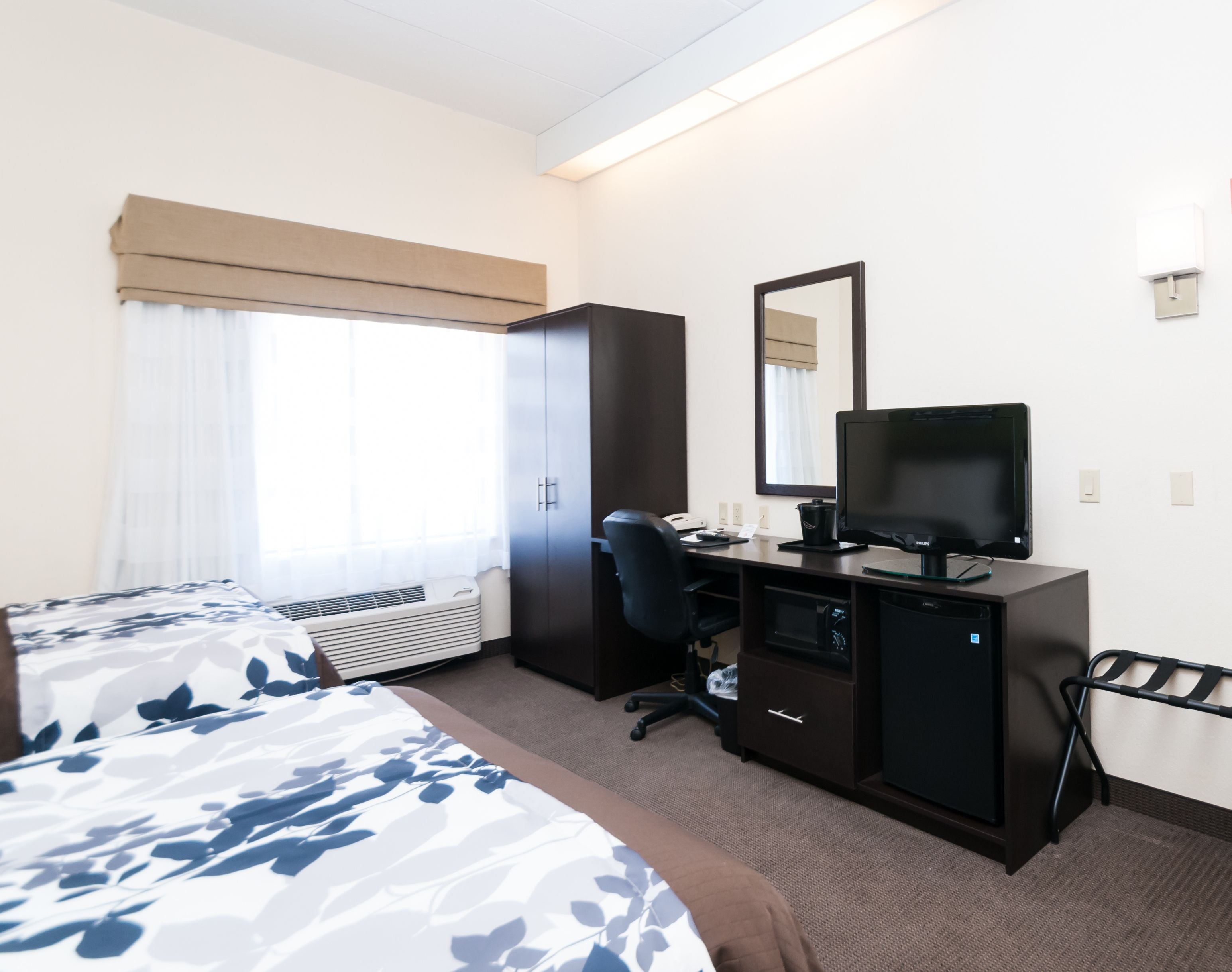Habitación de hotel con cama, escritorio con ordenador, silla, armario y ventana grande con cortinas.