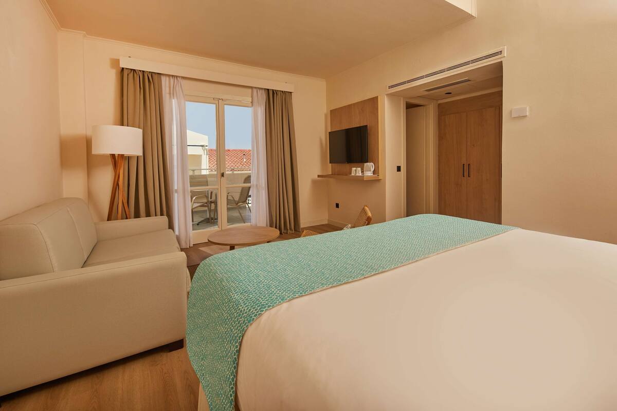A hotel room with a bed, a sofa, a small table, a TV, and a sliding door to a balcony.
