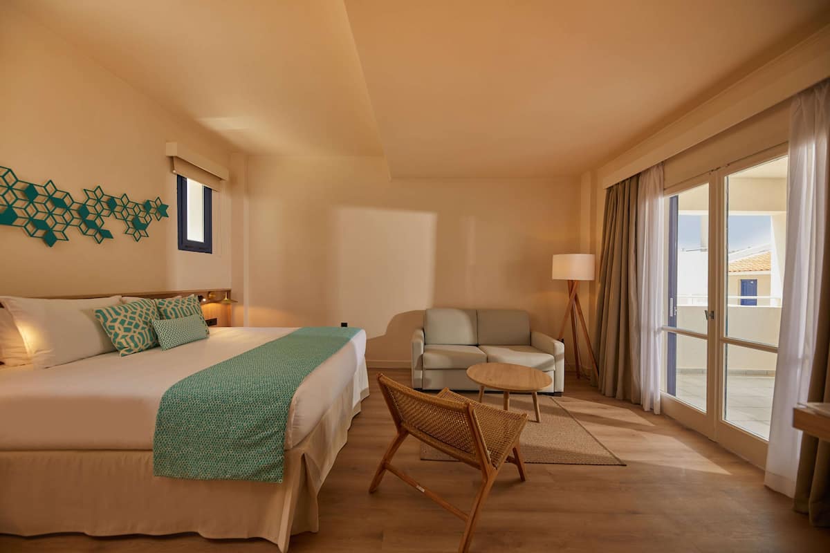 A hotel room with a bed, a sofa, a small table, and a chair. There is a window with curtains and a lamp.
