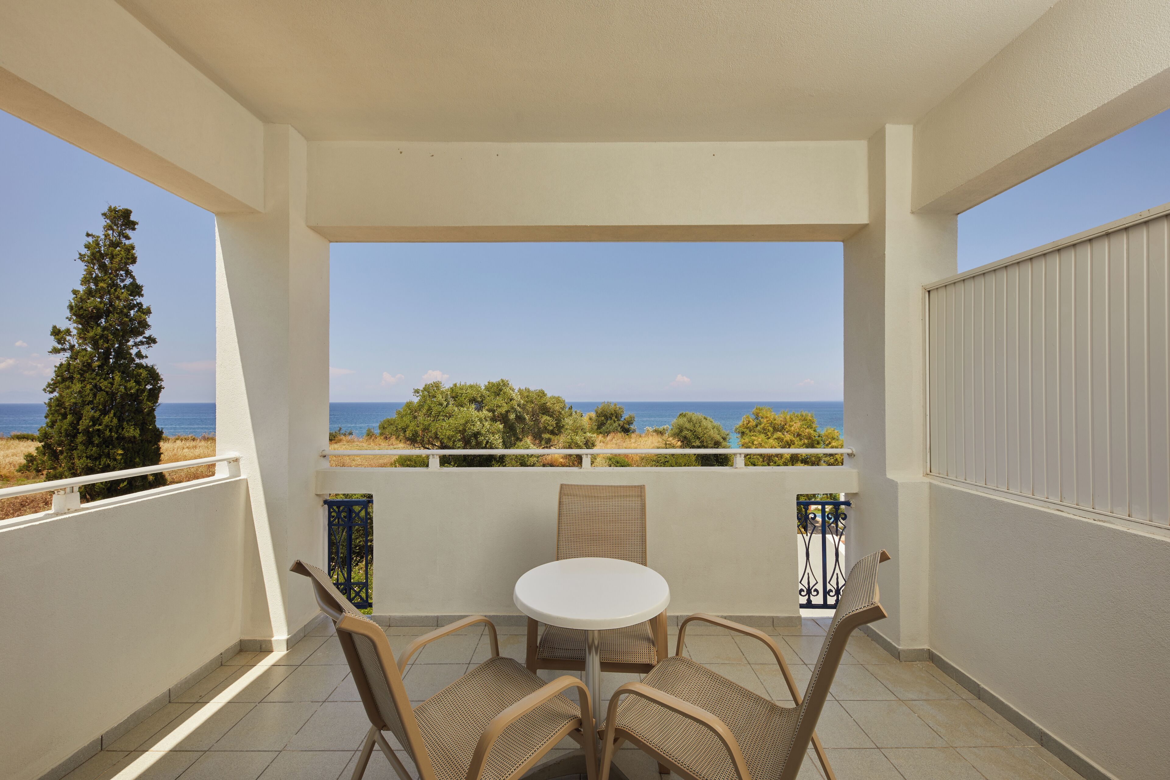 Un balcon avec une table blanche et des chaises en osier, donnant sur un paysage côtier avec des arbres et la mer.
