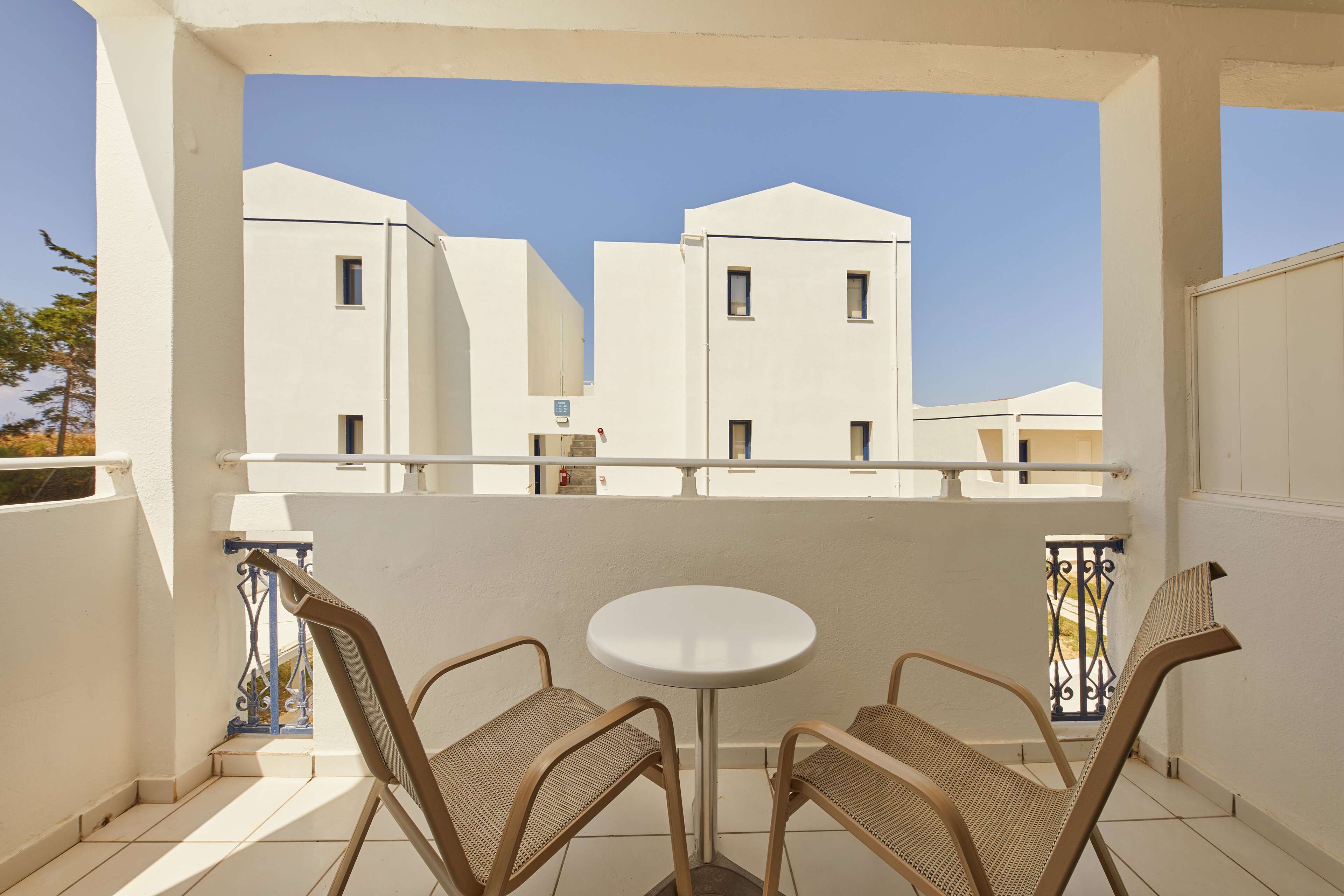 A balcony with two wicker chairs and a small round table, overlooking white buildings with multiple windows.