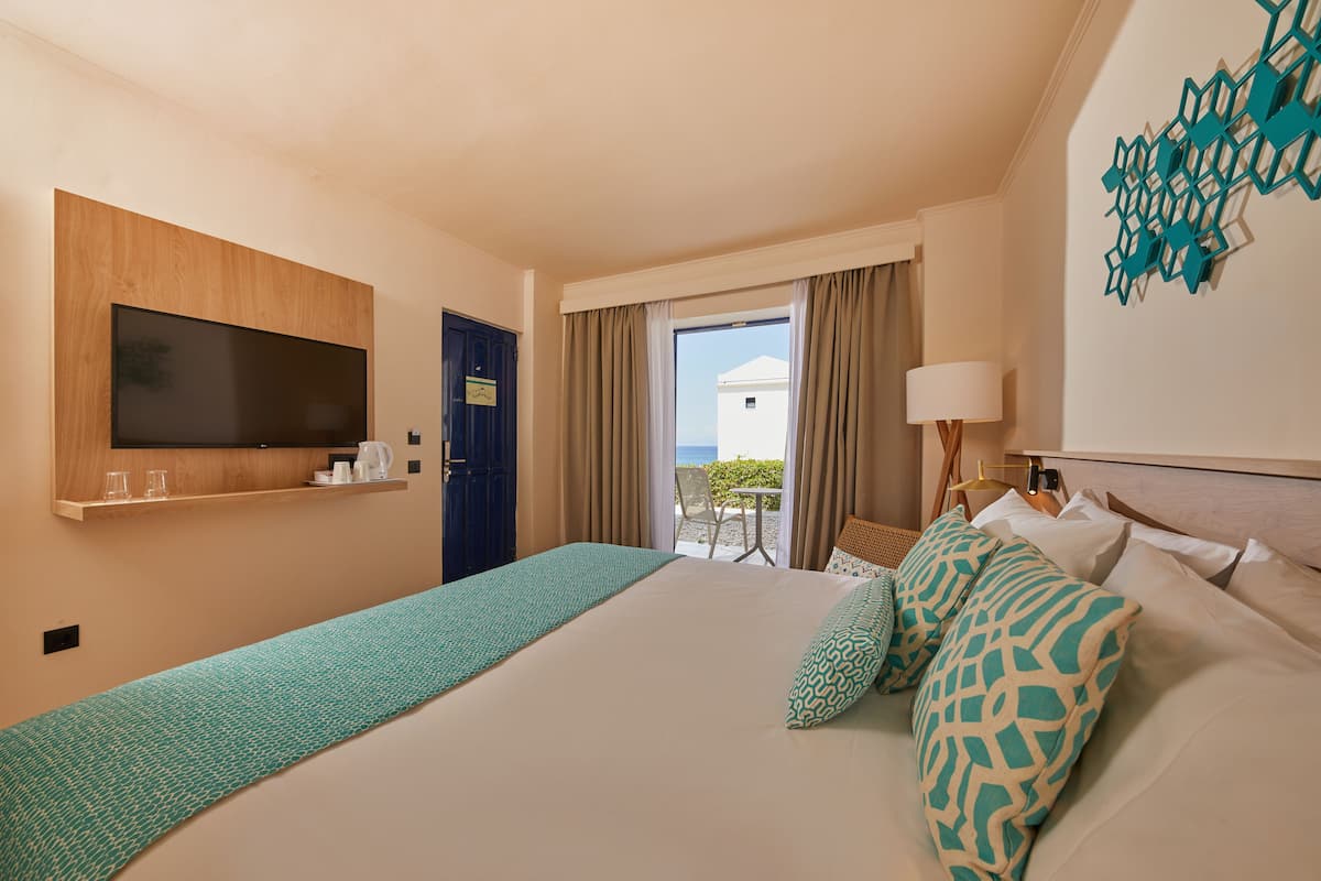 A hotel room with a large bed, a TV mounted on the wall, a balcony with a table and chairs, and a view of the sea.