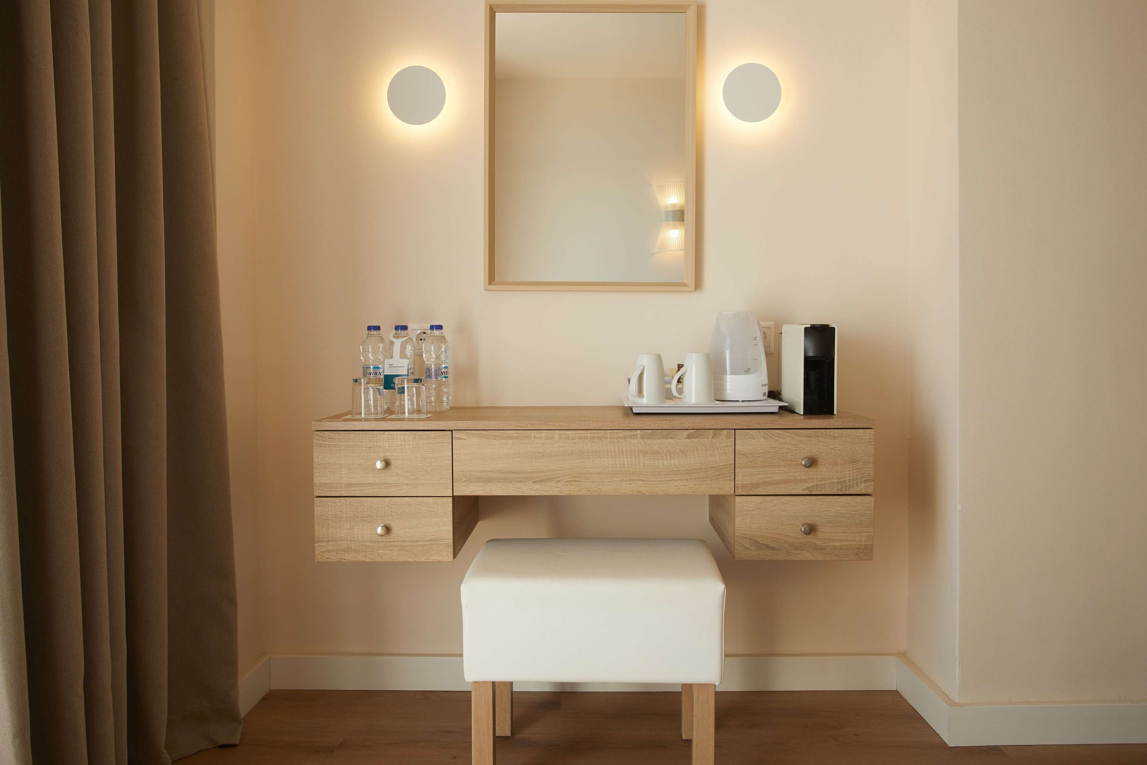 A hotel room with a wooden desk, a white bench, a mirror with wall sconces, and a coffee maker.