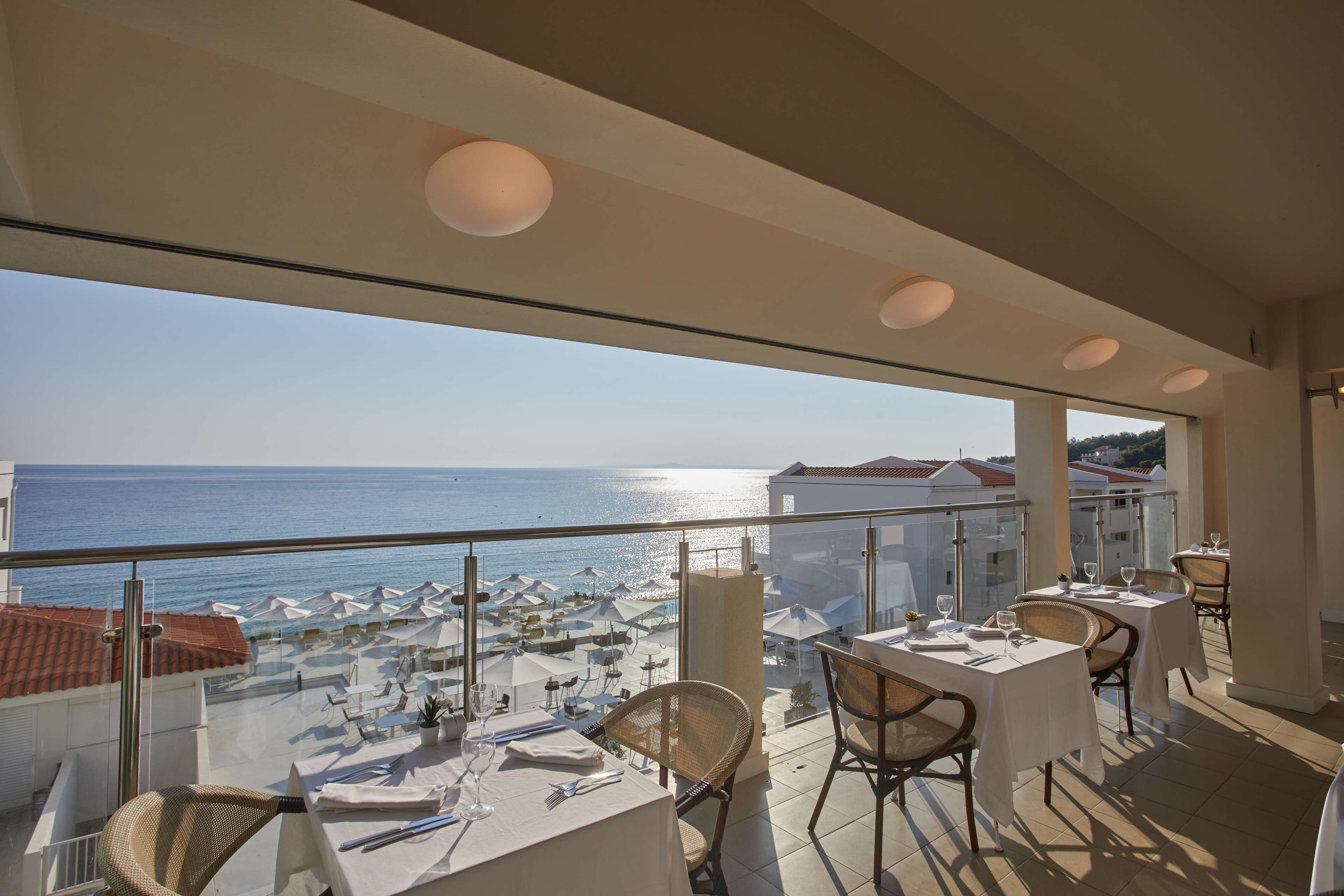 Une terrasse avec des tables et des chaises, donnant sur une plage parsemée de parasols et sur la mer.