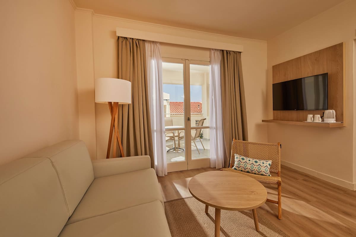 A hotel room with a sofa, a round table, a wicker chair, a TV mounted on the wall, and a sliding glass door leading to an outdoor area.