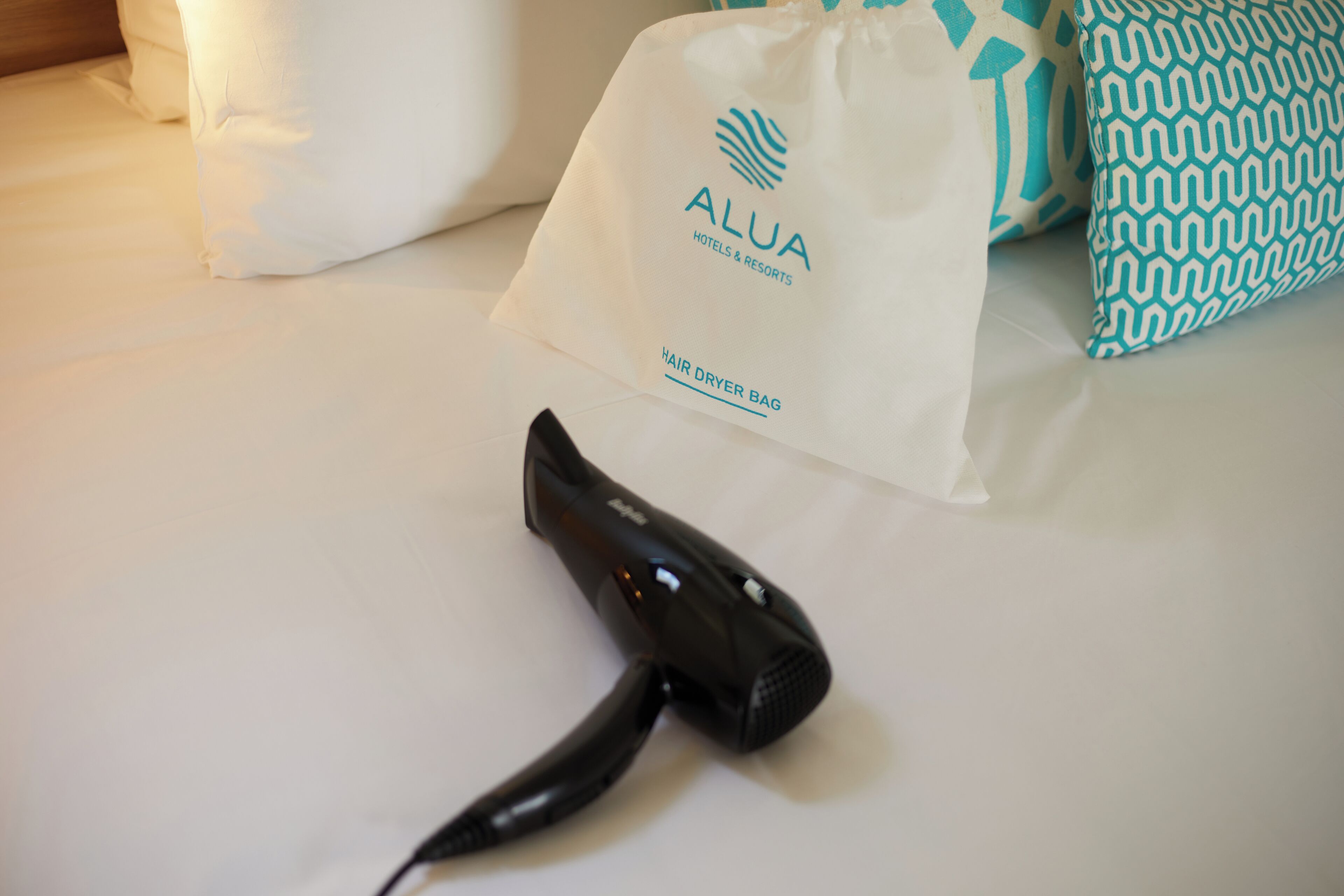 Un sèche-cheveux posé sur le lit, un oreiller blanc et un sac blanc orné du logo « ALUA HOTELS & RESORTS » et de l’inscription « SAC POUR SÈCHE-CHEVEUX ».