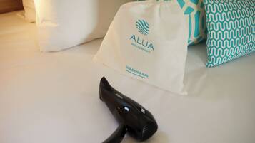 Un sèche-cheveux posé sur le lit, un oreiller blanc et un sac blanc orné du logo « ALUA HOTELS & RESORTS » et de l’inscription « SAC POUR SÈCHE-CHEVEUX ».