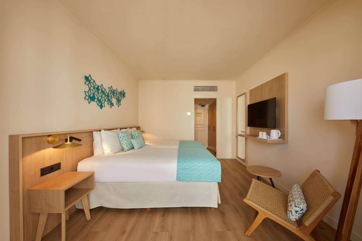 A hotel room with a bed, bedside table, lamp, chair, TV, and a small table with a cup and saucer.