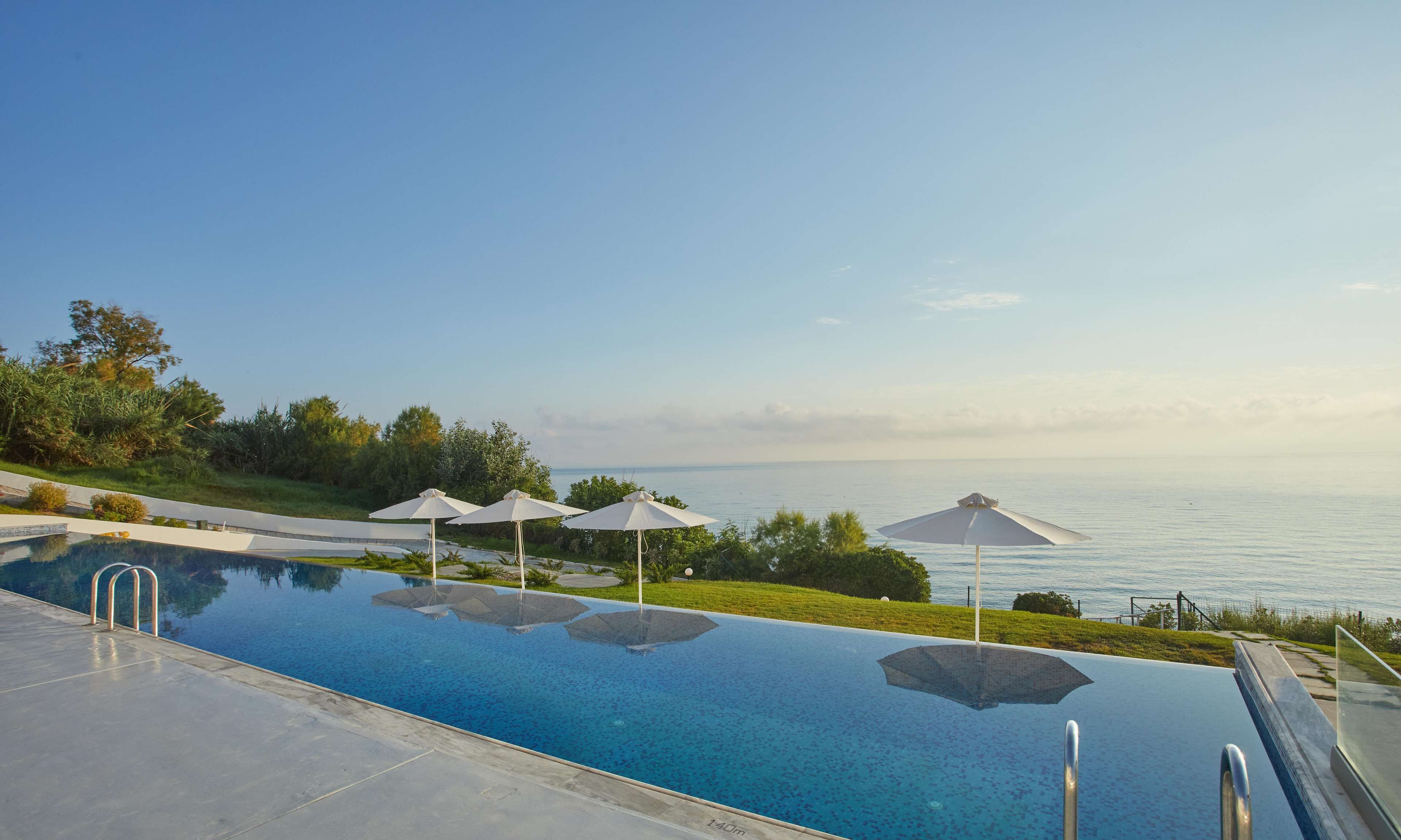 A swimming pool with a view of the sea, surrounded by greenery and umbrellas.