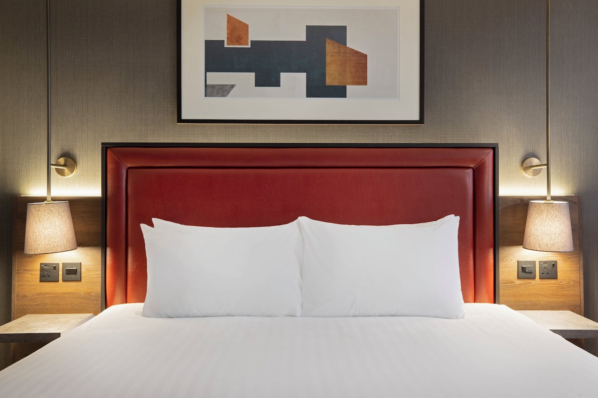 Quarto de hotel com cabeceira estofada vermelha, roupa de cama branca e duas luminárias laterais.