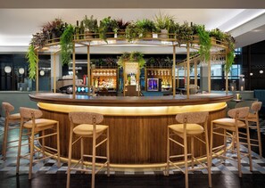 A bar area with a wooden counter, bar stools, and a variety of plants. - Leonardo Royal Hotel Birmingham  (Birmingham)
