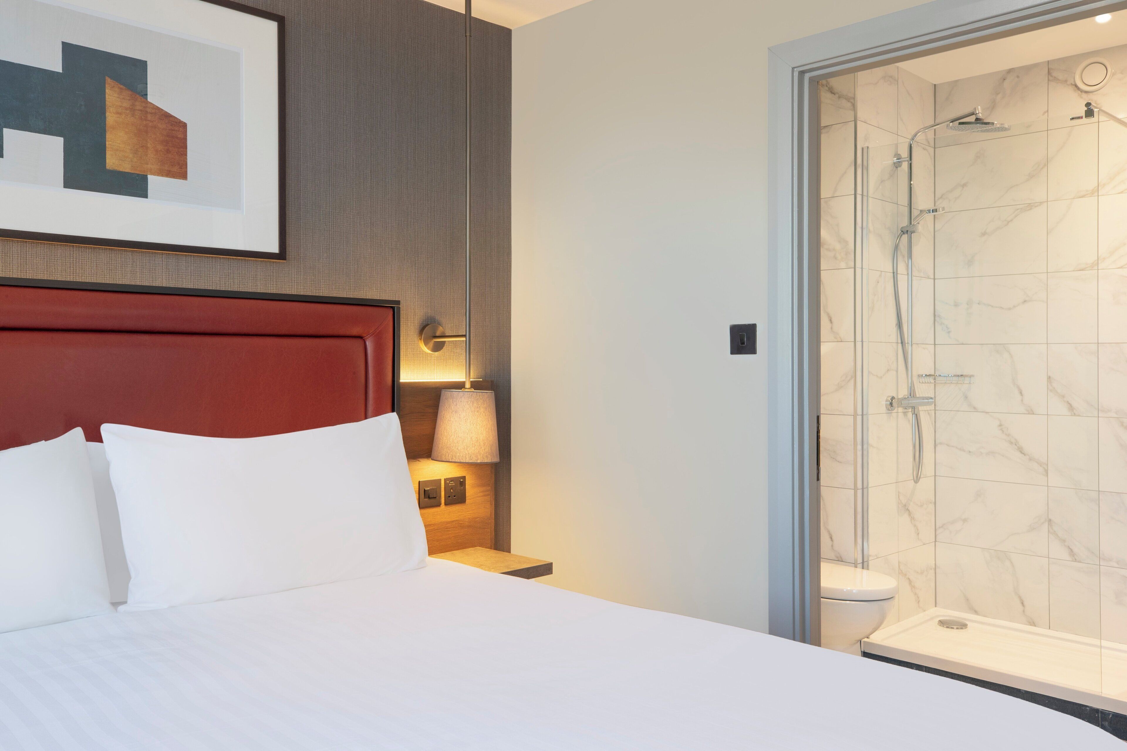 Een hotelkamer met een bed, een nachtkastje met lamp, een douche met glazen deur en een lijst met een afbeelding aan de muur.