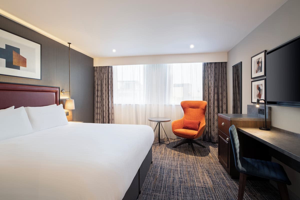 Quarto de hotel com cama grande, escrivaninha, cadeira laranja, mesa pequena e televisão.