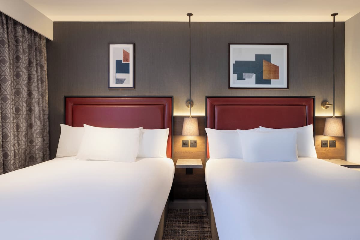Quarto de hotel com duas camas, cada uma com cabeceira de madeira e roupa de cama branca. Há luminárias fixadas na parede, de cada lado das camas, e duas obras de arte abstratas emolduradas sobre elas.