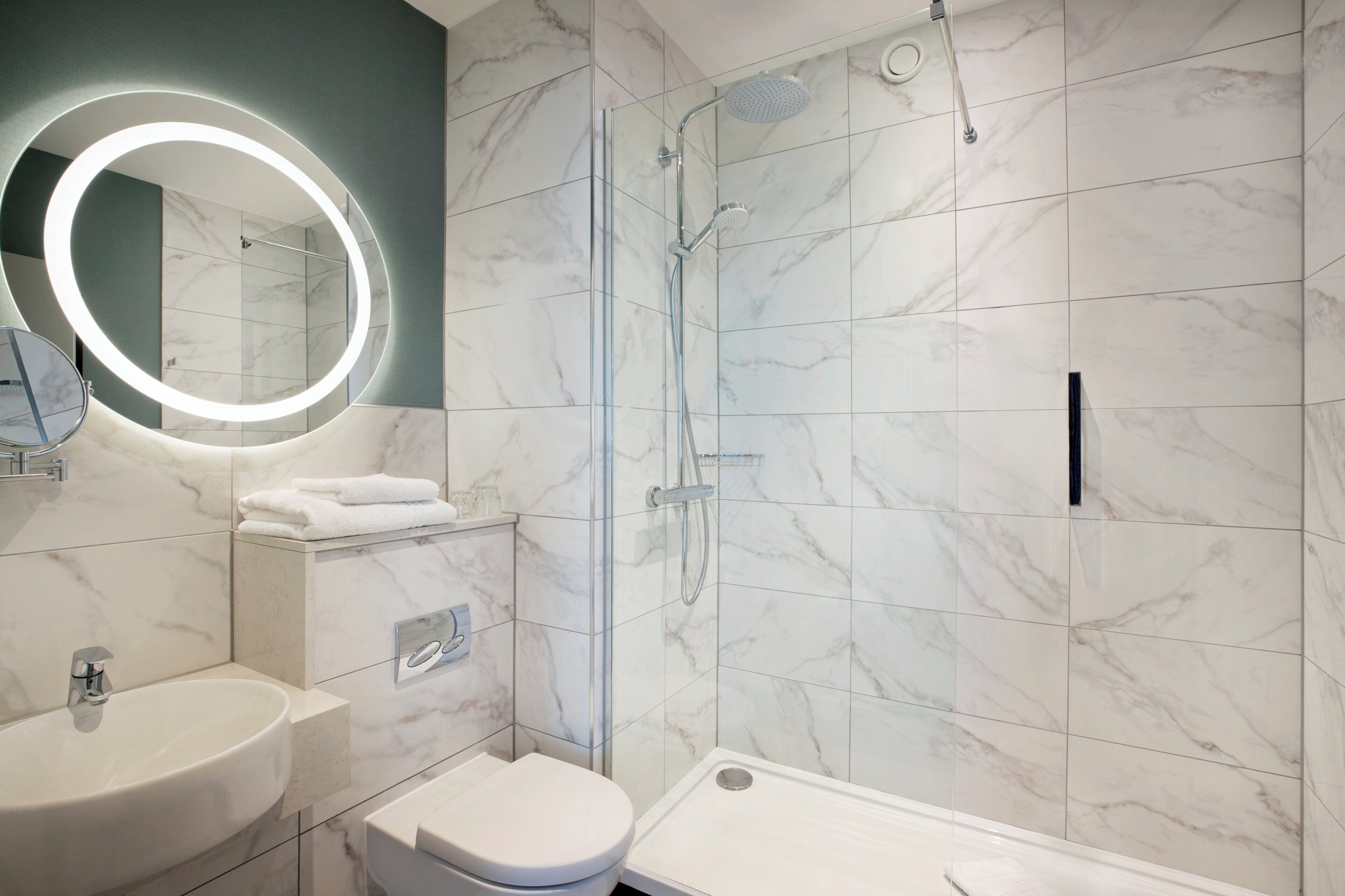 Un baño moderno con ducha de vidrio, un espejo redondo con luces LED, un lavamanos blanco y un inodoro.