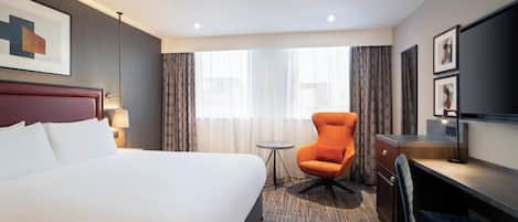 A hotel room with a large bed, a desk, an orange chair, a small table, and a TV.