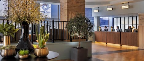 A modern hotel lobby with a reception desk, a large potted tree, and various indoor plants.