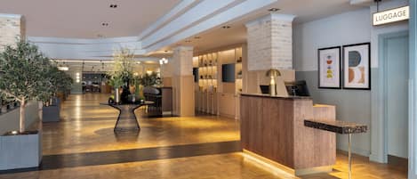 Un vestíbulo de hotel con mostrador de recepción, cartel de equipaje y un diseño moderno con suelo de madera y plantas en macetas.
