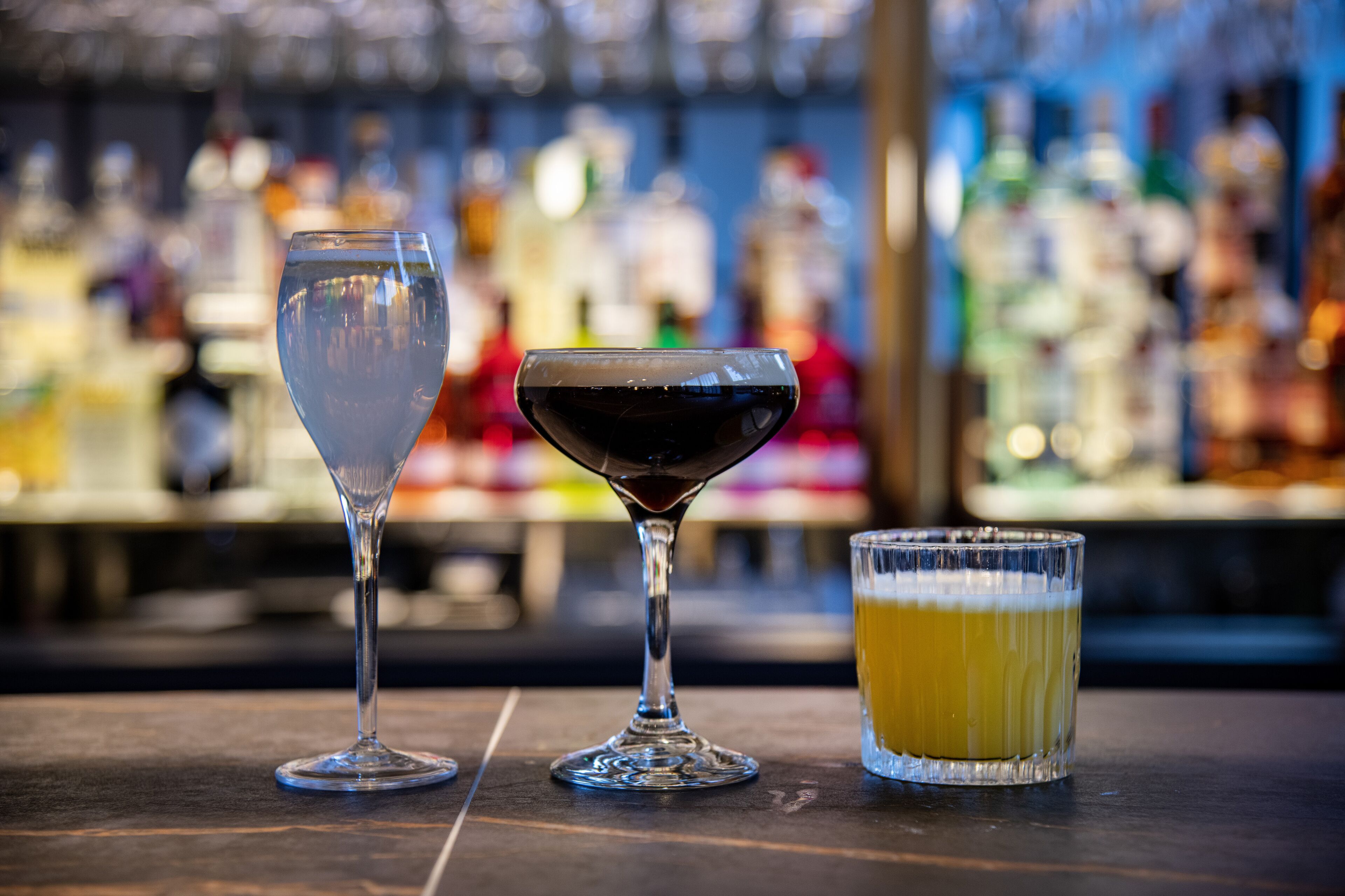 Tres vasos de bebidas distintas sobre una barra, con un fondo borroso de botellas.