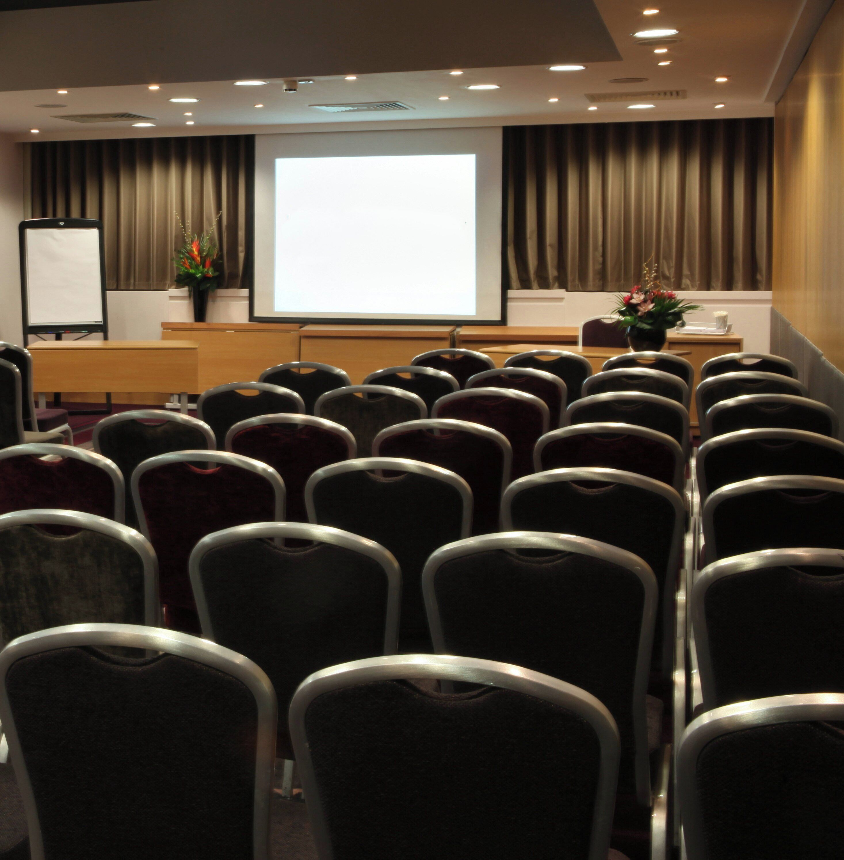 A conference room with rows of chairs facing a projection screen, a whiteboard, and a desk with a plant.