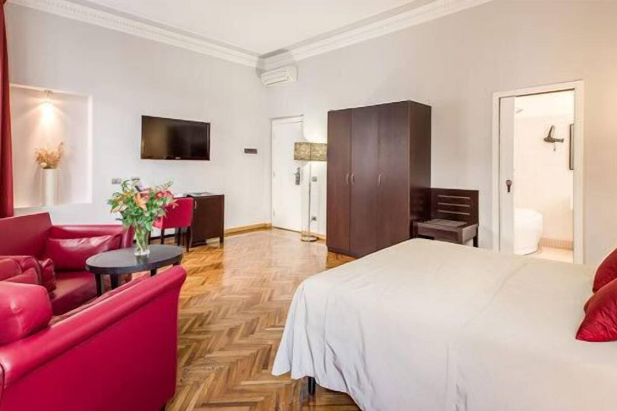 A hotel room with a bed, red sofa, a desk with a chair, a TV mounted on the wall, and a wardrobe.