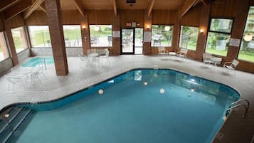 An indoor swimming pool with clear glass walls, surrounded by white chairs and tables, and a view of the outdoors.