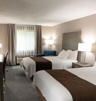 Quarto de hotel com duas camas, uma escrivaninha, uma cadeira, um abajur e uma janela com cortinas.