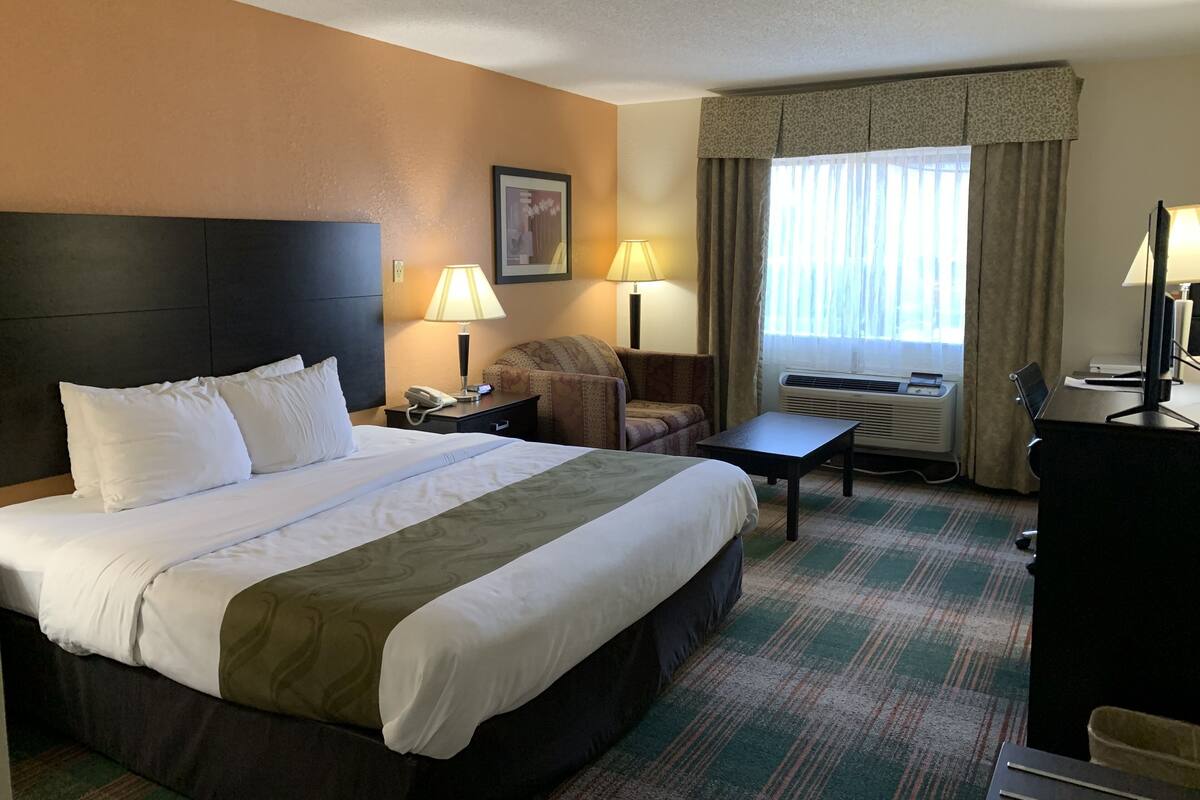 Une chambre d’hôtel avec un grand lit, un bureau, une chaise, deux lampes, un tableau encadré au mur et une fenêtre avec des rideaux.