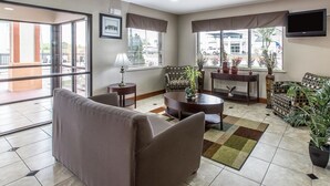 A modern living room with a sofa, coffee table, and patterned chairs. There are large windows with curtains, a flat-screen TV mounted on the wall, and a potted plant. - Quality Inn Montgomery South (Montgomery)