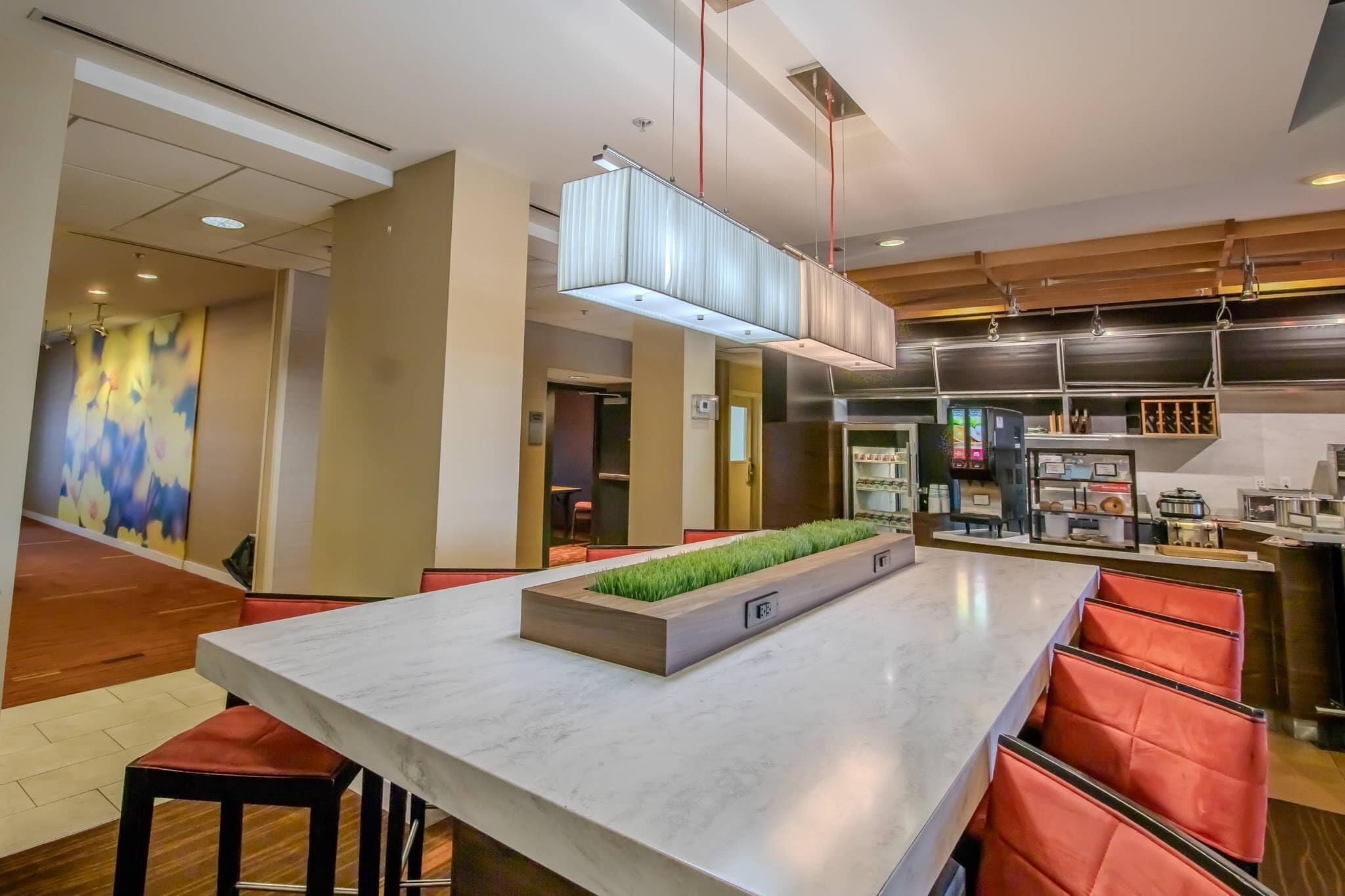 A modern hotel lobby with a marble reception desk, red cushioned seating, a decorative planter with green plants, a large illuminated light fixture, and a kitchen area in the background.