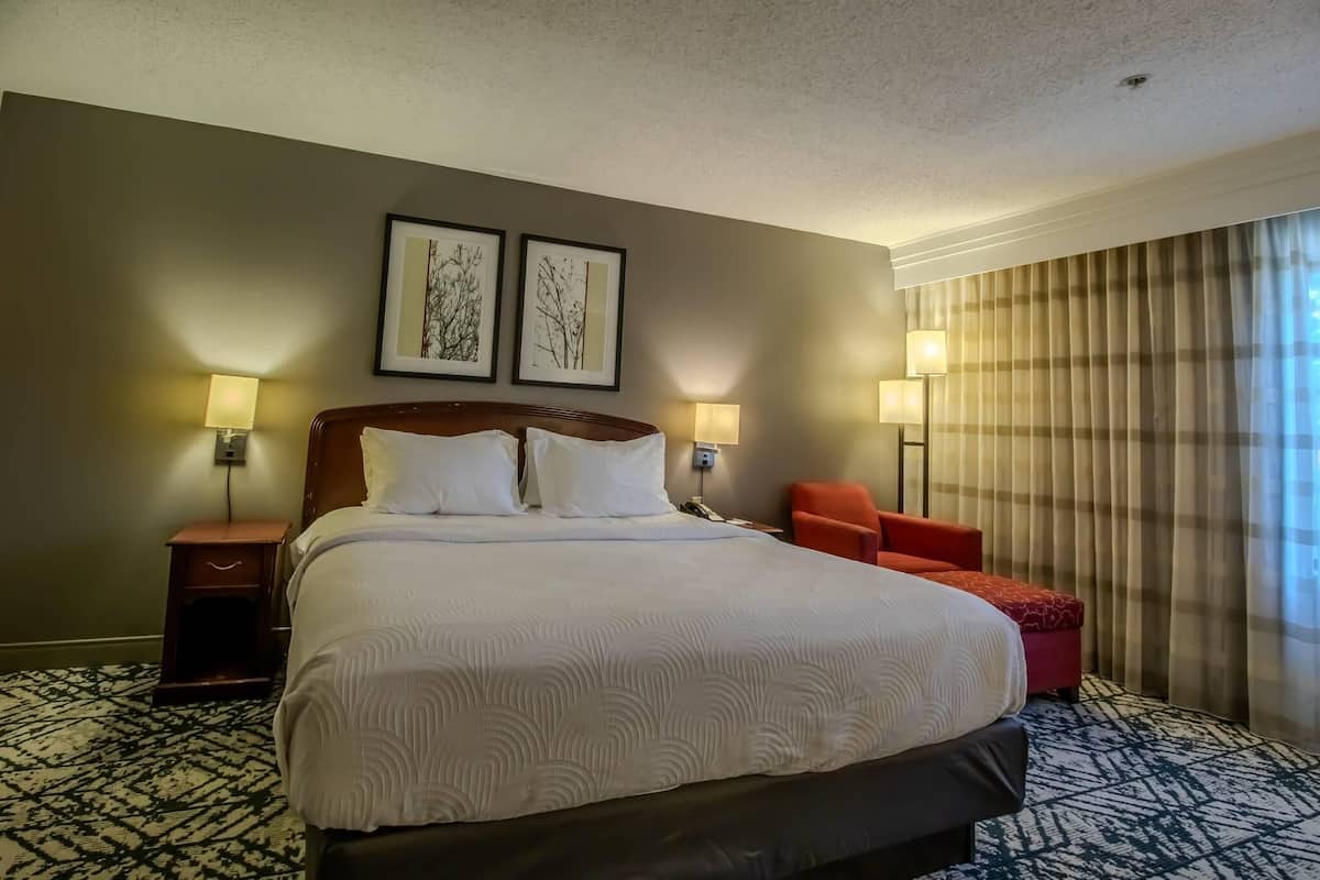 A hotel room with a large bed, two bedside lamps, a red chair, and a window with curtains.