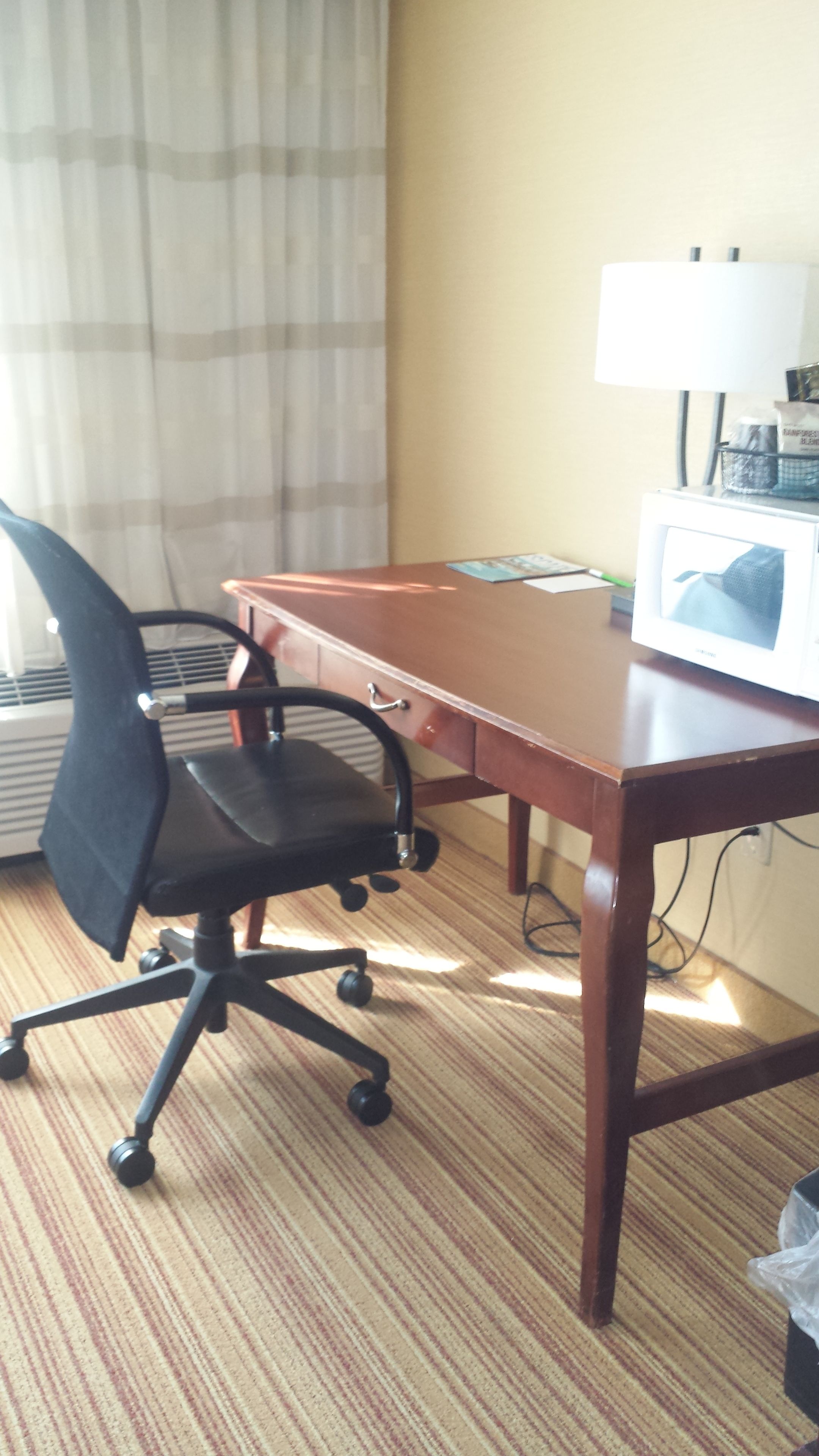 A hotel room with a wooden desk, a black office chair, a television, and a window with curtains.