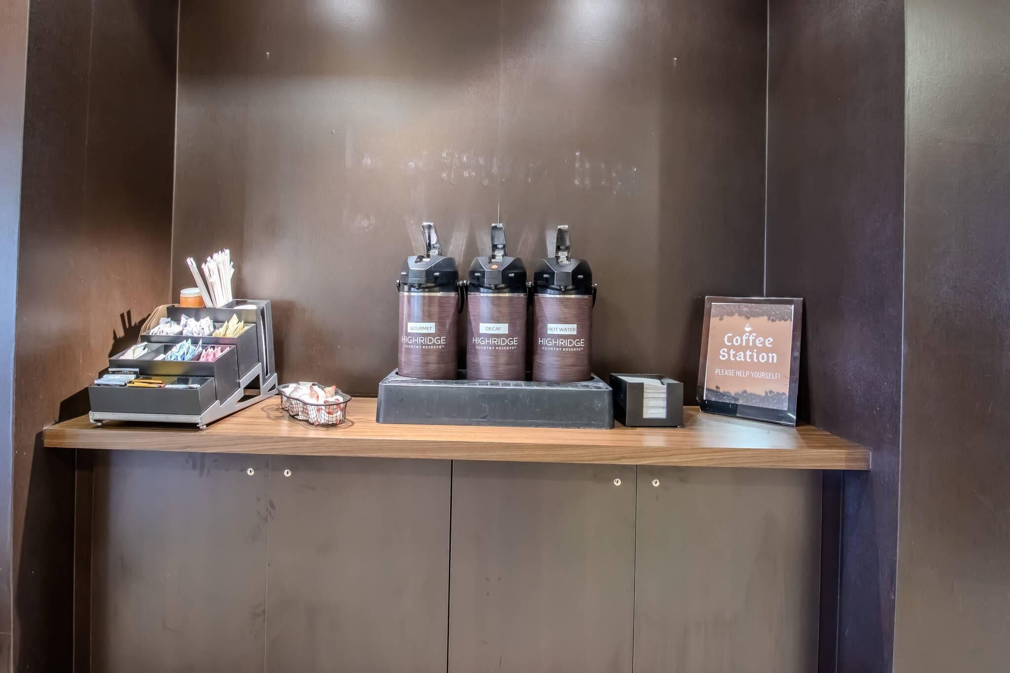 Estación de café con dispensadores etiquetados 'Gourmet', 'Descafeinado' y 'Agua Caliente', una máquina de café y un cartel que dice 'Estación de café, ¡sírvase usted!'