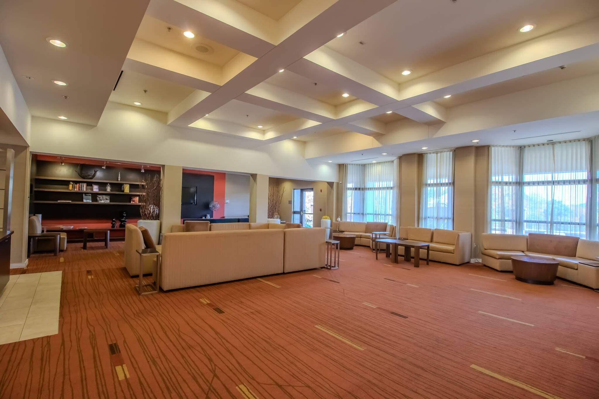 A spacious hotel lobby with a large sofa, multiple seating areas, a bookshelf, and large windows.