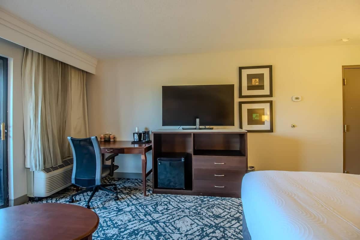A hotel room with a bed, a desk with a chair, a television, a small refrigerator, and a patterned carpet.