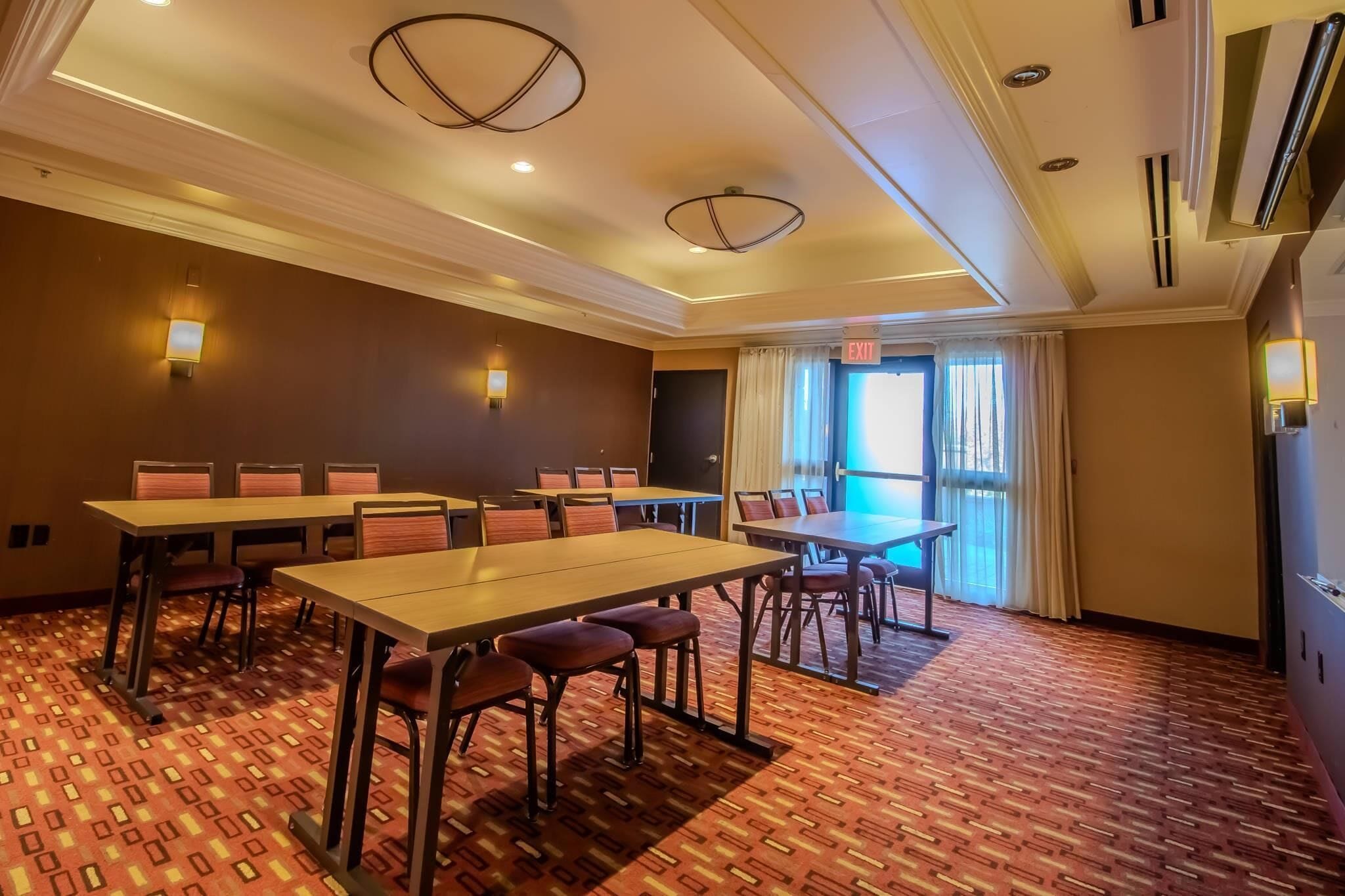 A conference room with tables and chairs, a patterned carpet, ceiling lights, and an exit sign.