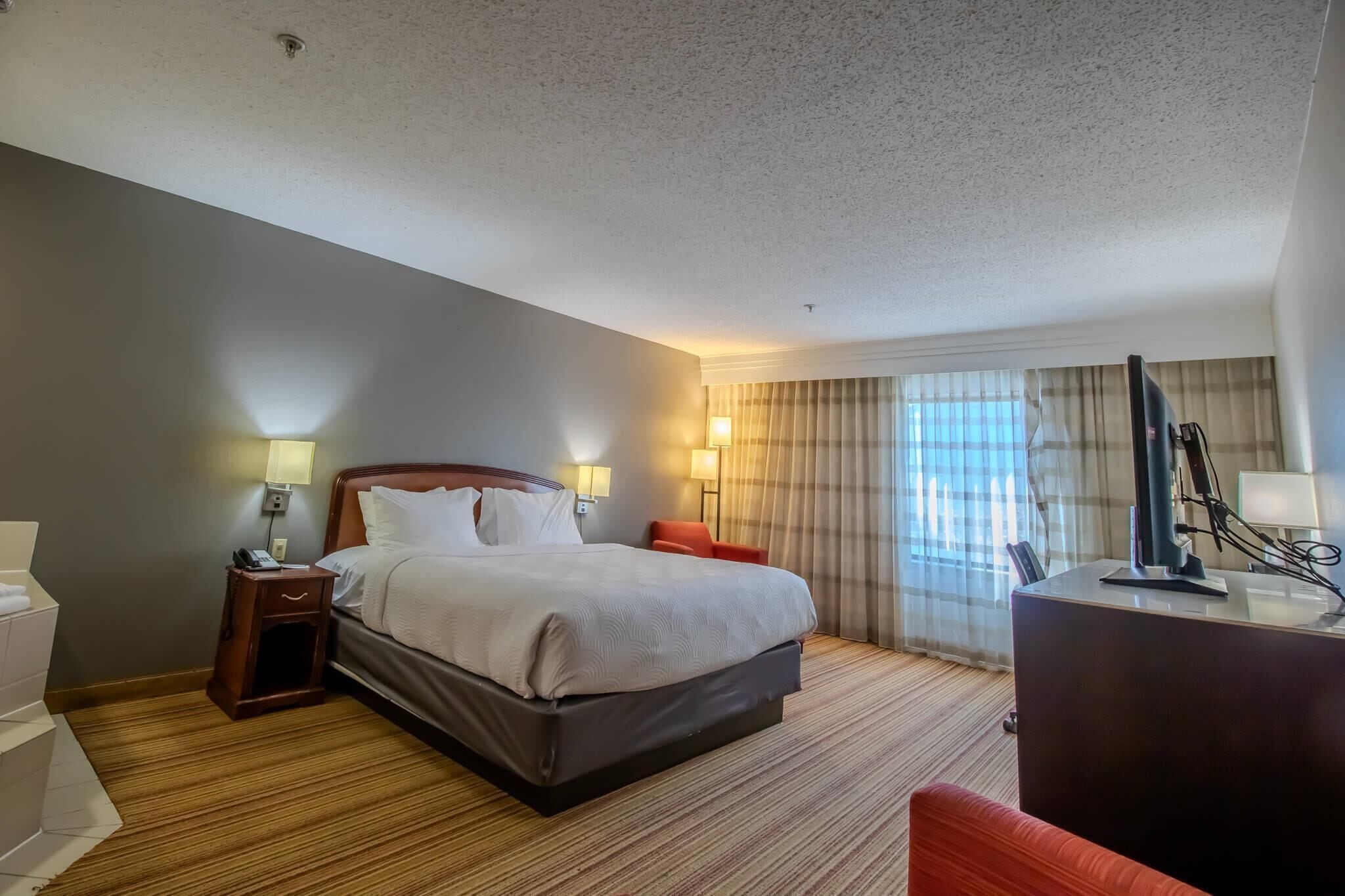 Habitación de hotel con una cama grande, lámparas de noche, un escritorio con una computadora y un ventanal con cortinas.