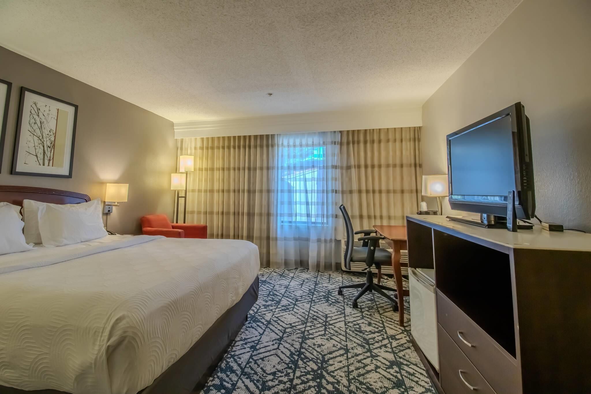 A hotel room with a large bed, a desk with a chair, a television, and two lamps.