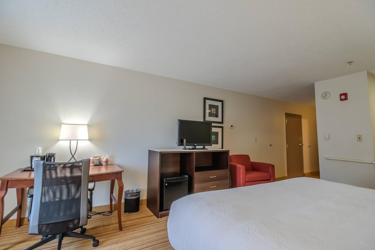 A hotel room with a bed, desk, chair, TV, and a red chair.