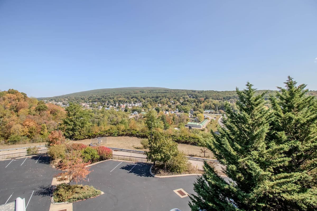 A parking lot with trees and a view of a town and hills in the distance.