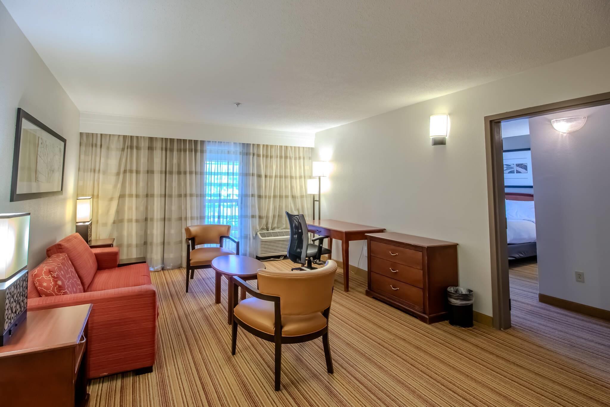 Habitación de hotel con sofá, sillón, escritorio y una mesita. Se ve una ventana con cortinas, una lámpara y un cuadro colgado en la pared.