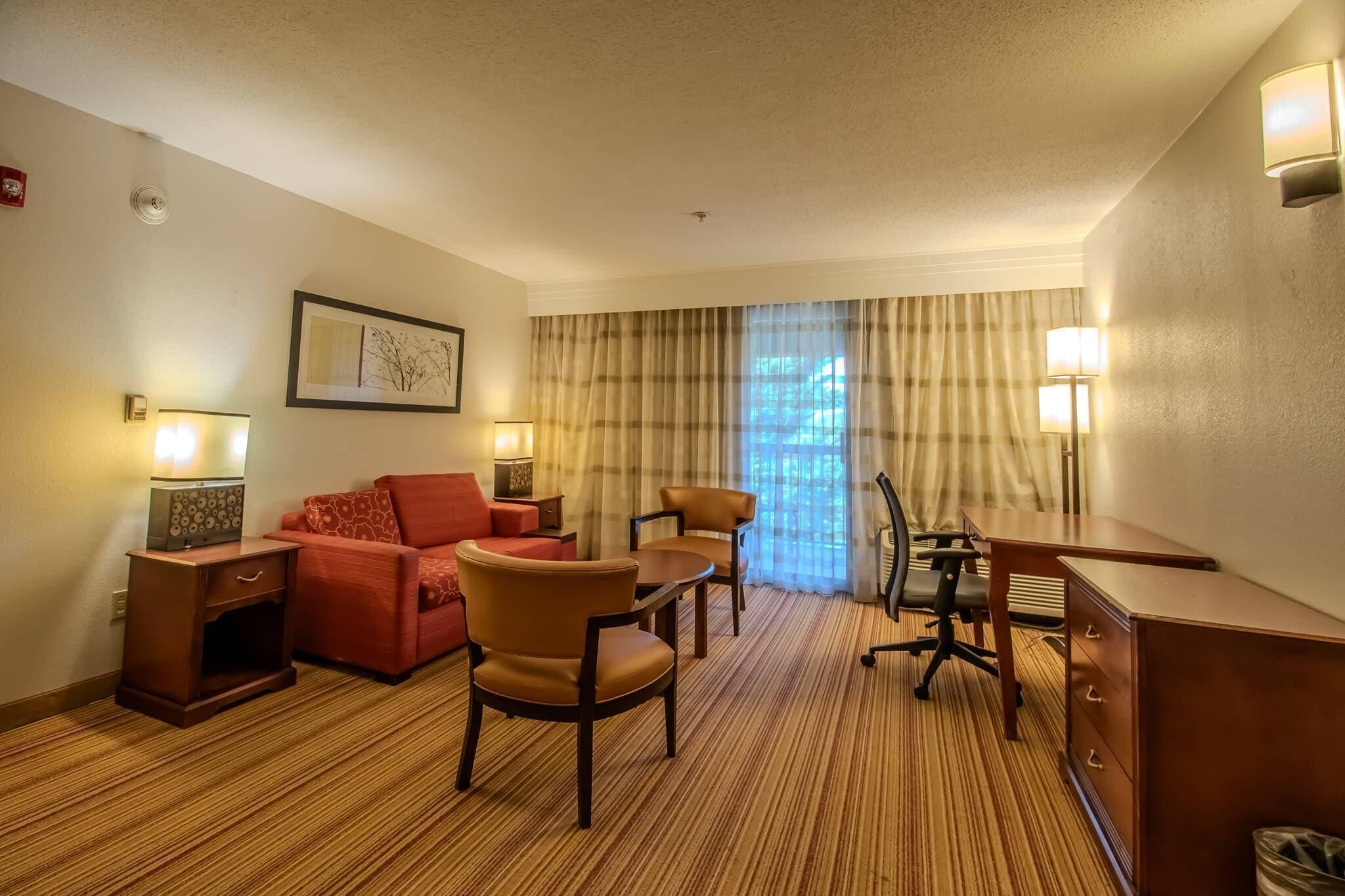 A hotel room with a red sofa, two armchairs, a small table, a desk, and a lamp.