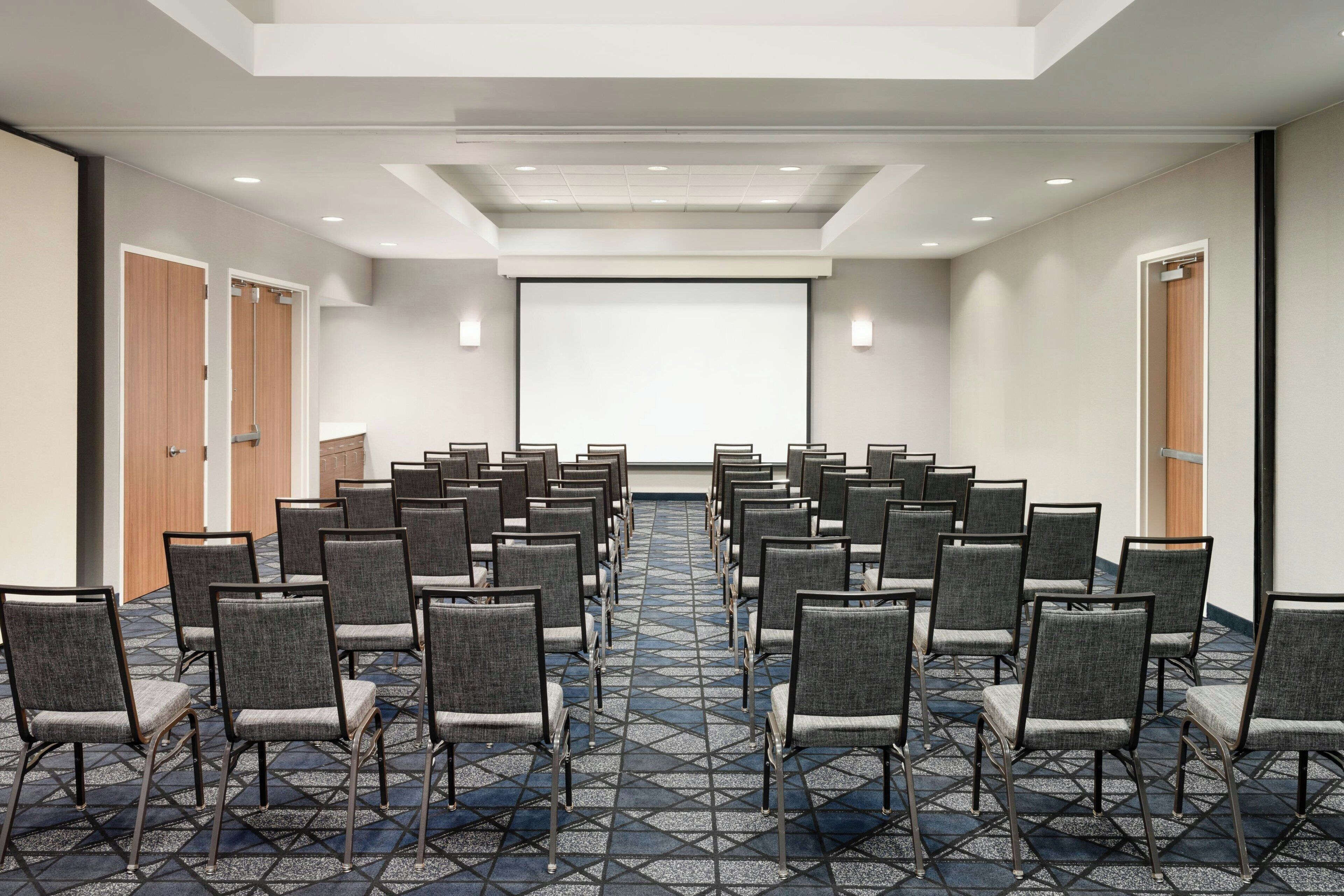 A conference room with rows of chairs facing a projection screen, carpeted floor, and doors on the sides.