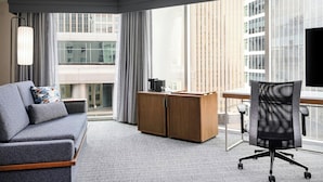 Una habitación de hotel moderna con sofá gris, escritorio de madera, silla de oficina negra y grandes ventanales con vistas a la ciudad.