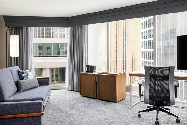 A modern hotel room with a grey sofa, a wooden desk, a black office chair, and large windows offering a city view. - Courtyard by Marriott Charlotte City Center (Charlotte)